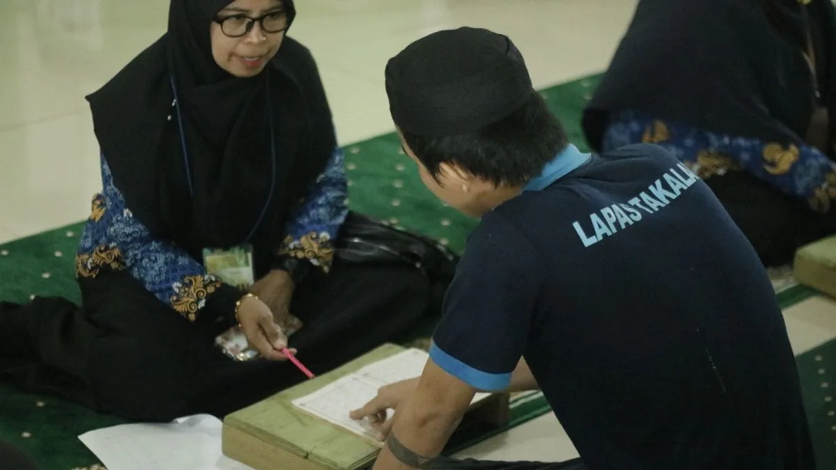 Seorang petugas penyuluh agama wanita berkacamata sedang membimbing seorang warga binaan pria memakai kopiah hitam belajar membaca Al-Qur'an di atas karpet masjid Lapas Takalar